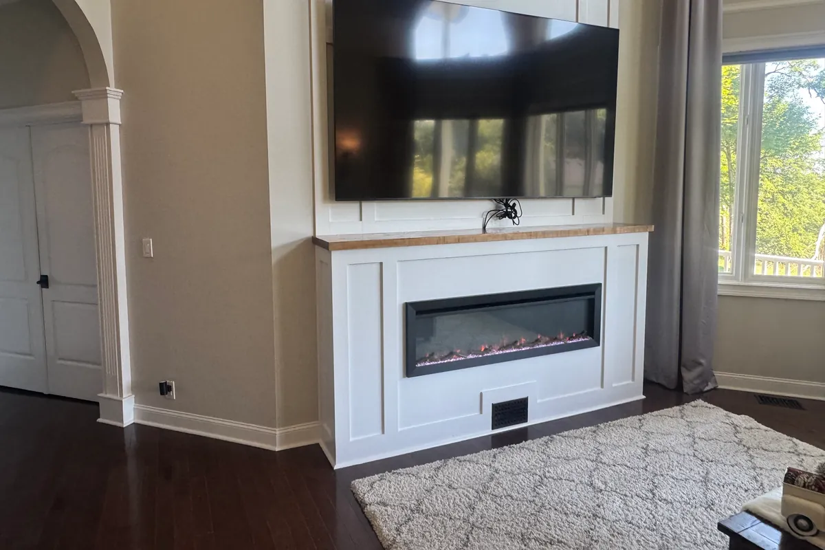 Wall-mounted flat screen TV centered above a linear electric fireplace, set into a white painted trim surround.