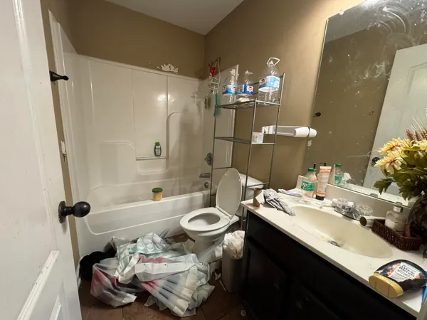 Bathroom left cluttered, with a wire shelf unit over the toilet holding water jugs, toiletries across the vanity and a shower curtain pulled down onto the floor.