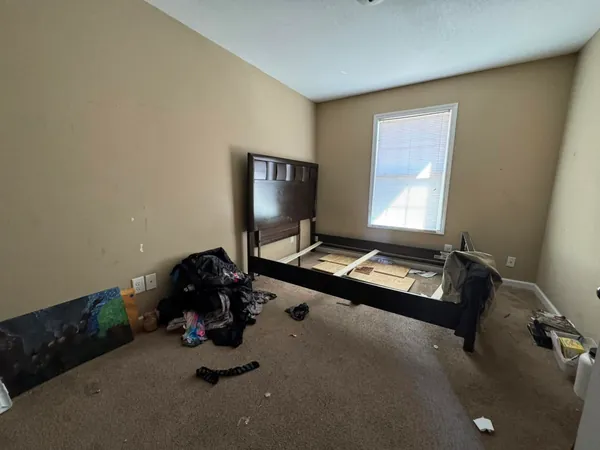 Bedroom with a bed frame taken apart and leaning against the wall, piles of clothing on the carpet and debris left across the floor.