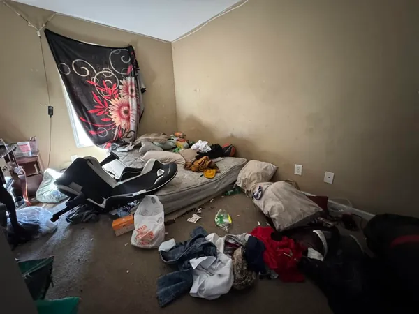 Bedroom left full after a move-out, with a mattress on the floor, a broken gaming chair on its side, and clothing, bedding and trash spread across the carpet.