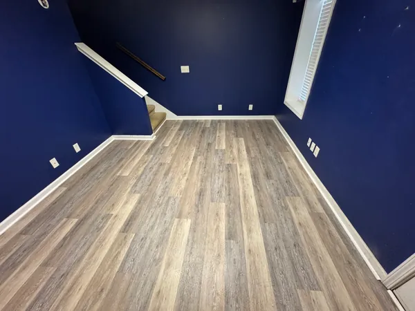 Empty room with new gray-brown luxury vinyl plank run wall to wall, white baseboard cut in around it, deep navy walls, a louvered closet door and a stair rail at the far corner.