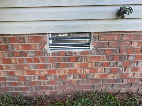 New aluminium foundation vent set into a red brick crawlspace wall and mortared in, with fresh mortar still light against the older joints and an outside tap on the siding above.