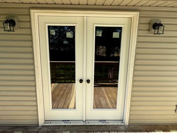 New cream double French patio doors with full-length glass, set into a vinyl-sided wall under a porch roof, trimmed out square with a black coach lantern on each side and the deck reflected in the glass.