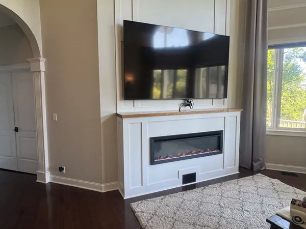 Wall-mounted flat screen TV centered above a linear electric fireplace, set into a white painted trim surround.