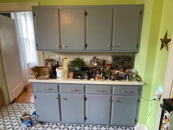The same kitchen cabinet run repainted a slate gray-blue with new brass bar pulls and knobs, the drawer and cabinet contents stacked out on the counter while the job finishes.