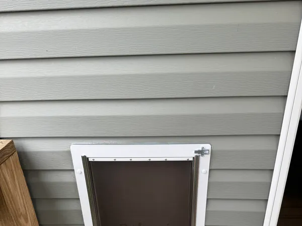 Pet door cut into a vinyl-sided exterior wall and trimmed out in white, with a brown flap and an aluminium frame, opening onto a wood deck beside a sliding door.