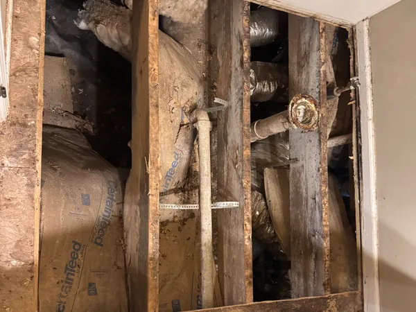 The same wall cut open to the stud bays, showing kraft-faced batt insulation, a cast iron drain stack, water lines and the back of the shower valve, with the cabinets still in place below.